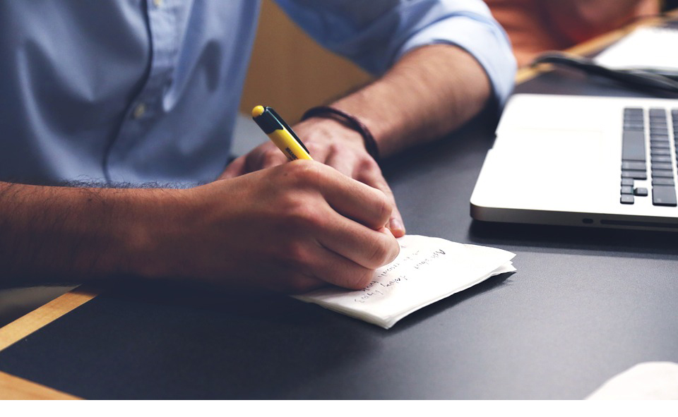 persona escribiendo en un papel frente a una computadora.