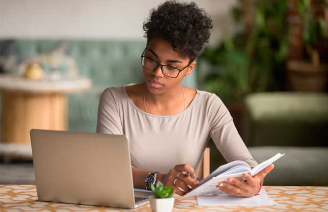 Mujer llevando a cabo un análisis de su investigación frente a la computadora, con el apoyo de un libro