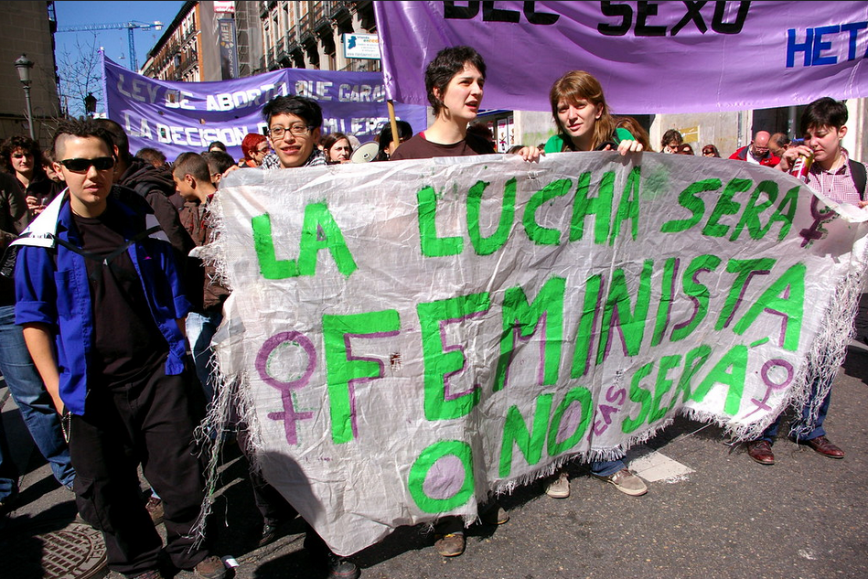 En una manifestación, cinco mujeres jóvenes sostienen pancarta que dice: La lucha será feminista o no será