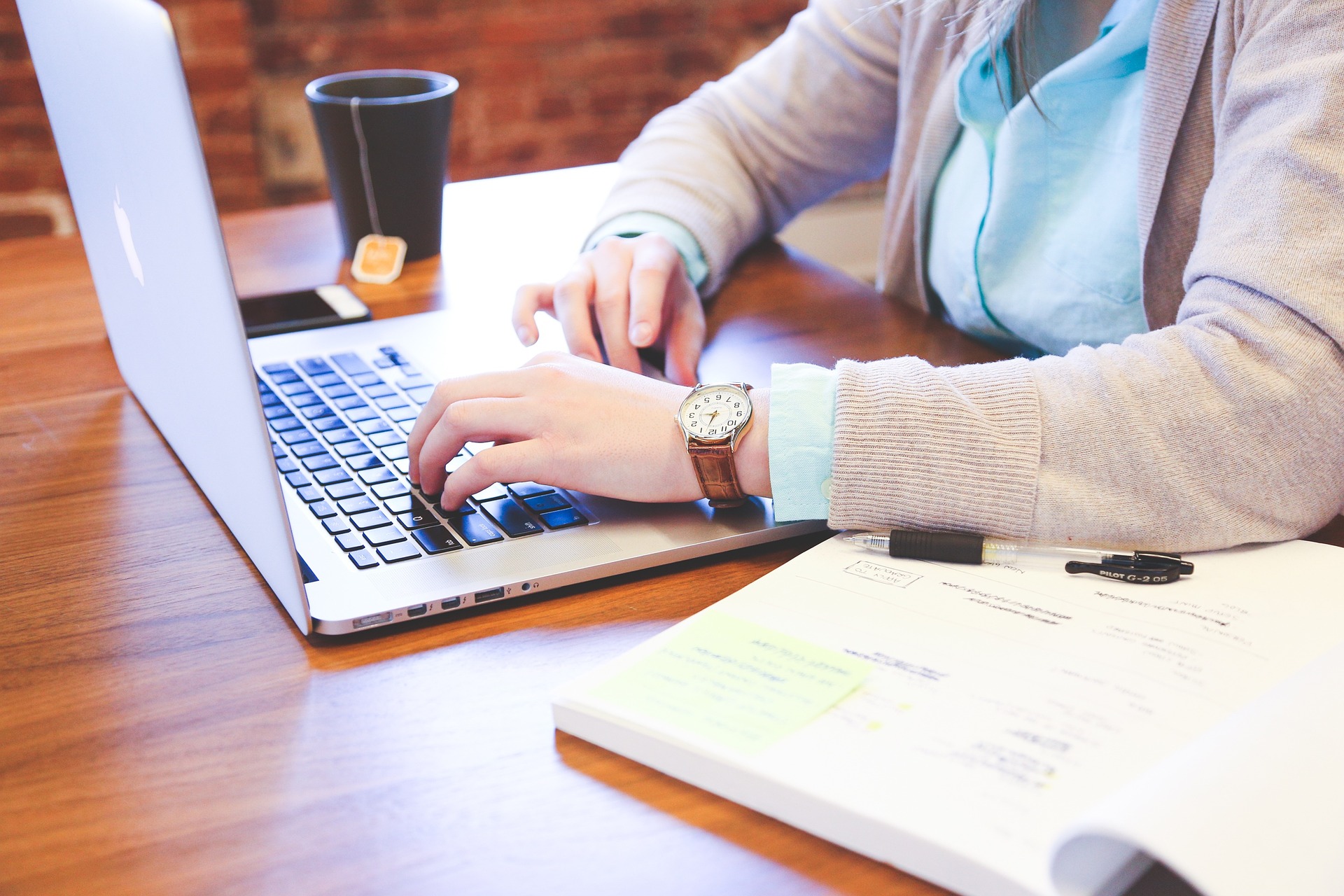 Torso de perfil de mujer sentada ante laptop, teclea y al fondo, sobre la mesa de madera, se ve un vaso de café y, al frente, un libro abierto.